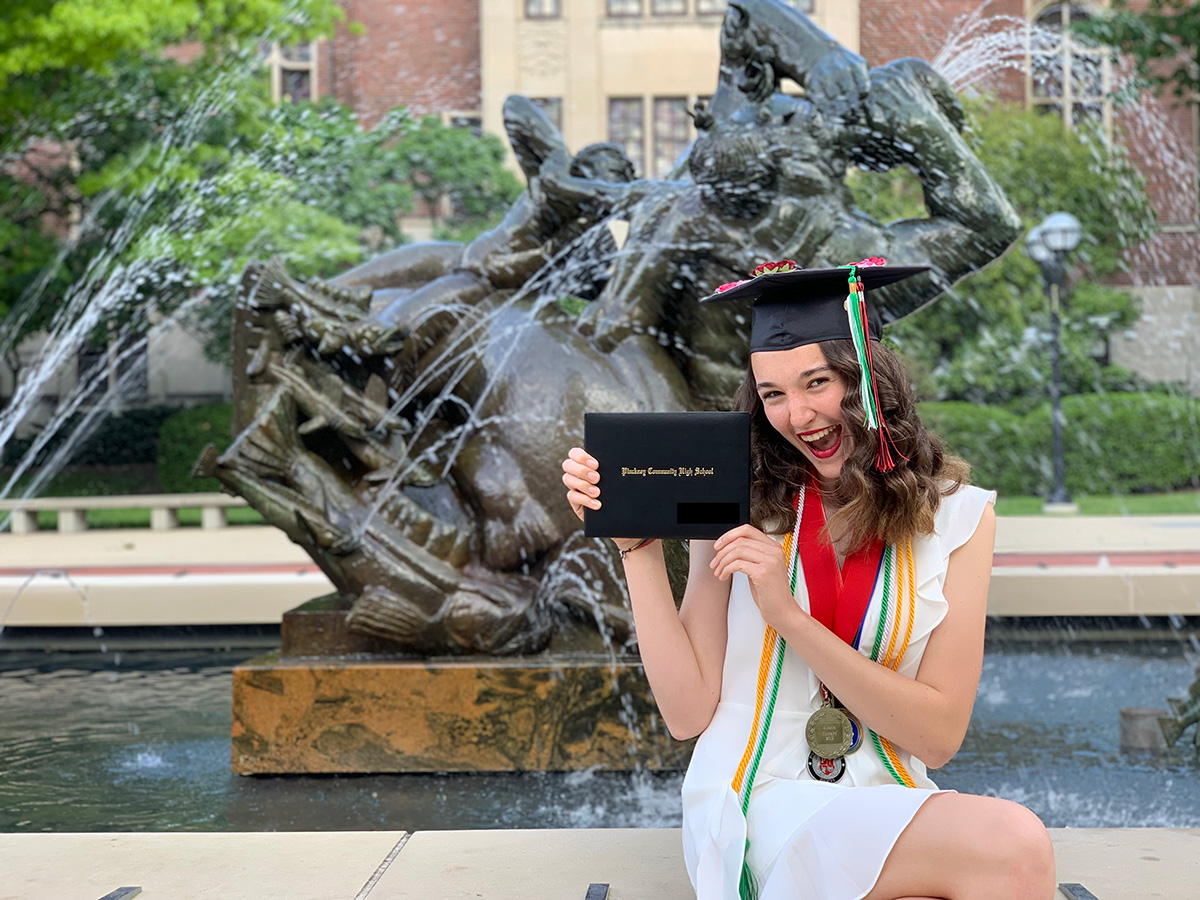 Photo of Alexis in front of campus fountain in high school graduation cap and gown holding diploma.
