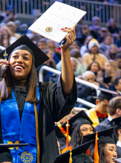 A graduate in a cap and gown talks on the phone while looking toward the crowd at Winter Commencement.