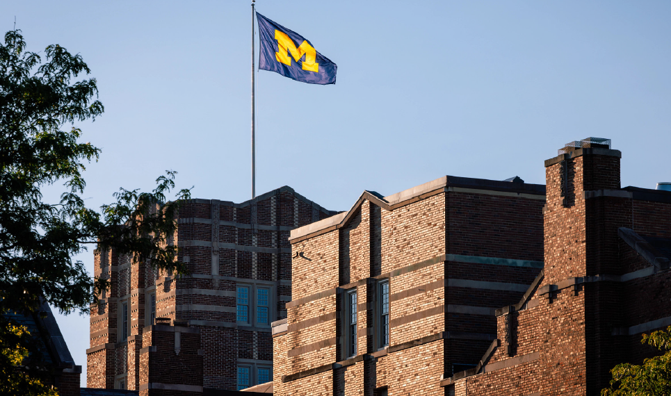 The Michigan Union flag waves against a clear blue sky during sunrise.