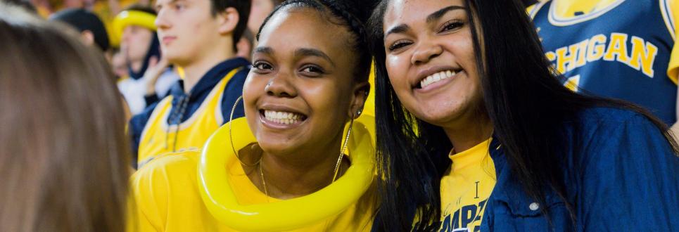 Two women students smiling while at a sporting event, wearing all maize and blue